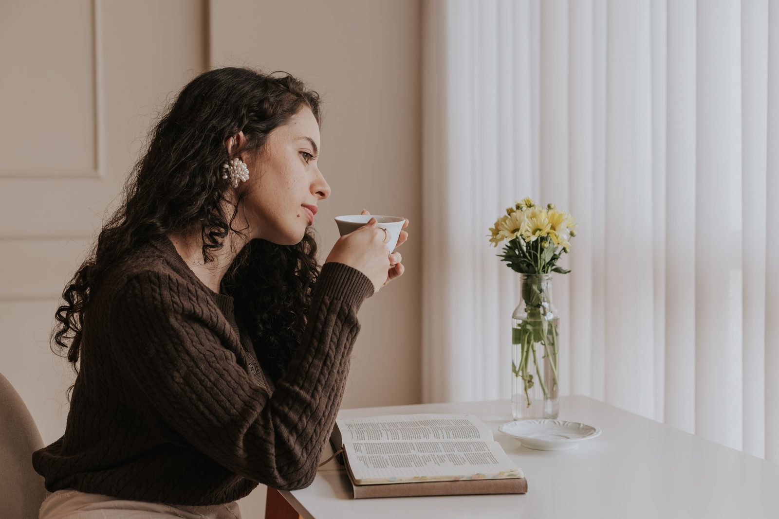 Mujer tomando café junto a la ventana, con su Biblia abierta y flores amarillas